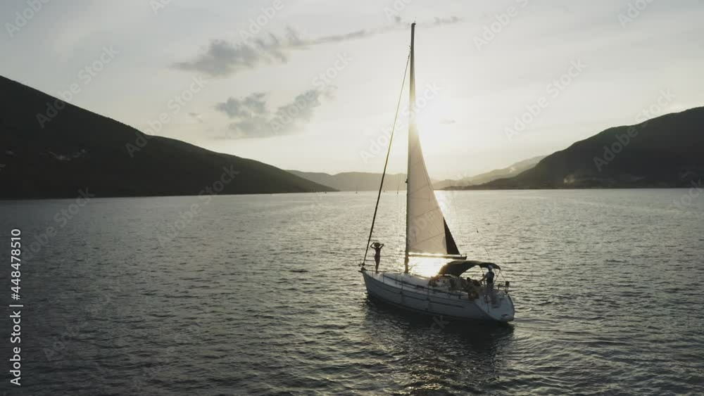 Evening romantic tracking shot of sailboat cruising on sea. Woman posing on bow deck. Water surface reflecting evening sun.