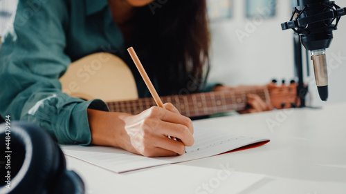 Happy asia woman songwriter play acoustic guitar listen song from smartphone think and write notes lyrics song in paper sit in living room at home studio. Music production at home concept.
