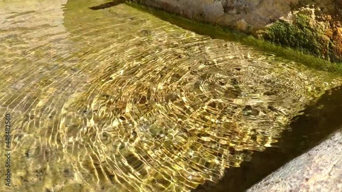 Close up of an stone fountain from a small town full of crystal clear water. It's very sunny and water drops falll creating circular ripples that reflect light on different tones. photo