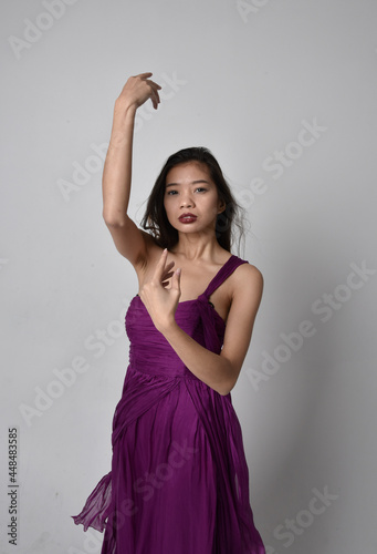 Close up portrait of pretty brunette asian girl wearing purple flowing gown. Gestural hand movements on grey studio background.