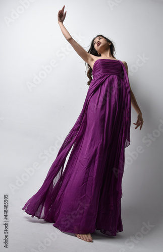 Full length portrait of pretty brunette asian girl wearing purple flowing gown. Standing pose in low angle on on studio background.