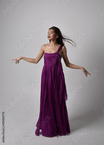 Full length portrait of pretty brunette asian girl wearing purple flowing gown. Standing, dancing pose on on studio background.