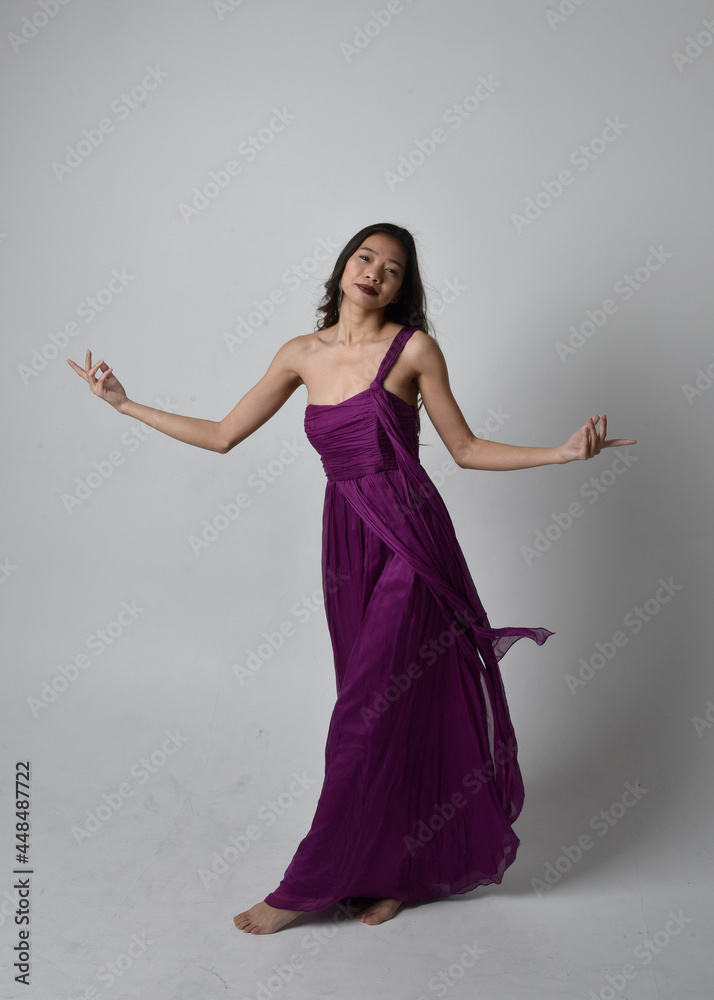 Naklejka premium Full length portrait of pretty brunette asian girl wearing purple flowing gown. Standing, dancing pose on on studio background.