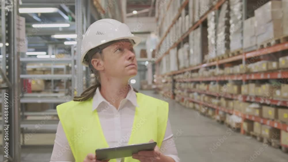 Woman manager or storekeeper in helmet walks through logistics warehouse using tablet to inventory control of goods. Woman manager or storekeeper in helmet walks through logistics warehouse using tablet to inventory control of goods.