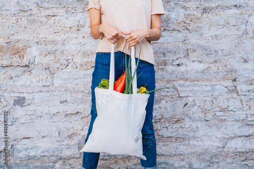 Woman holding white canvas tote bag with vegetables near brick wall.