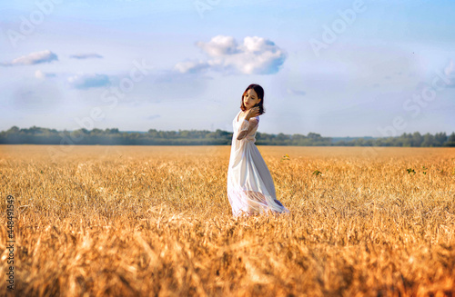 A young girl in a white long dress and a hat is standing in a wheat field