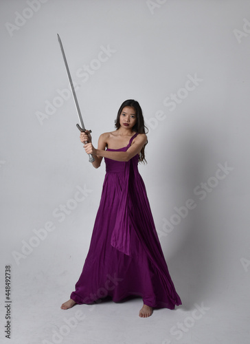 Full length portrait of pretty brunette asian girl wearing purple flowing gown. Sitting pose holding a sword on on studio background.
