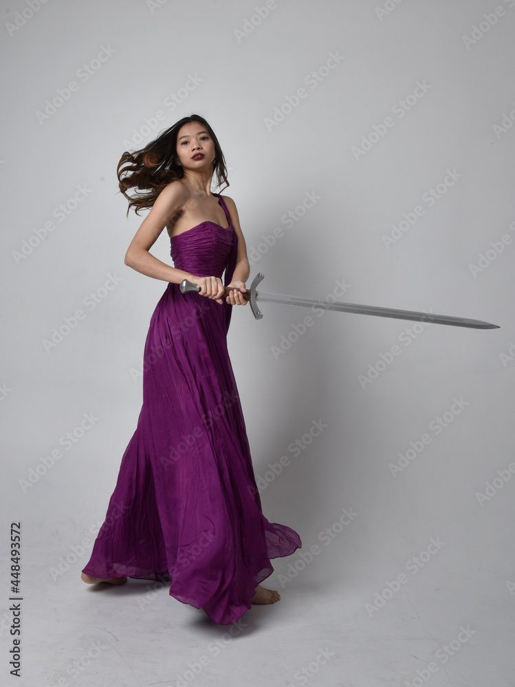 Full length portrait of pretty brunette asian girl wearing purple flowing gown. Sitting pose holding a sword on on studio background. Full length portrait of pretty brunette asian girl wearing purple flowing gown. Sitting pose holding a sword on on studio background.