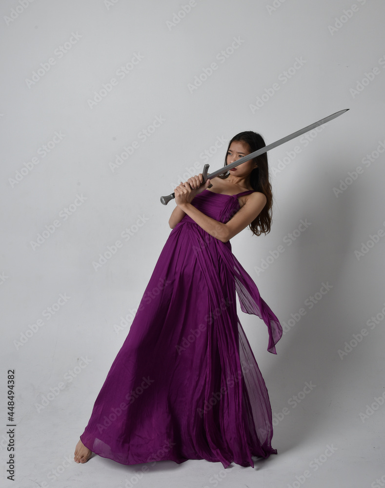 Full length portrait of pretty brunette asian girl wearing purple flowing gown. Sitting pose holding a sword on on studio background. Full length portrait of pretty brunette asian girl wearing purple flowing gown. Sitting pose holding a sword on on studio background.