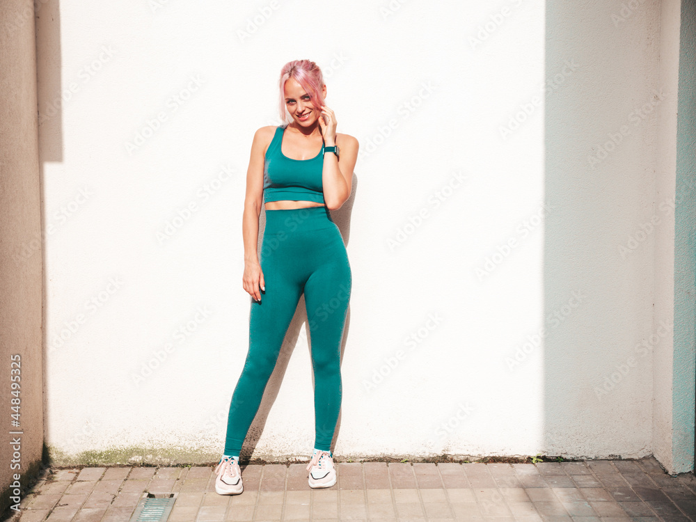 Fitness smiling woman in green sports clothing with pink hair. Young beautiful model with perfect body.Female posing in the street near white wall.Cheerful and happy. Stretching out before training Fitness smiling woman in green sports clothing with pink hair. Young beautiful model with perfect body.Female posing in the street near white wall.Cheerful and happy. Stretching out before training