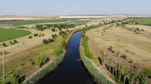 Aerial of an irrigation field canal constructed to convey water. Day drone view of Saratov area in Russia, around a field with green trees next to a dirt road.