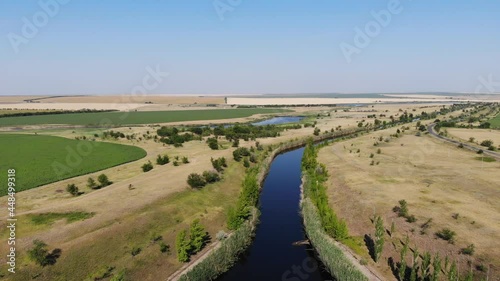 Aerial of an irrigation field canal constructed to convey water. Day drone view of Saratov area in Russia, around a field with green trees next to a dirt road.