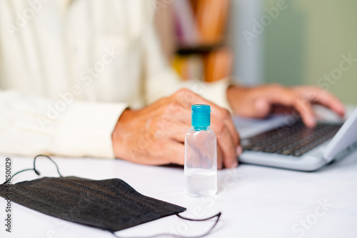 Focus on sanitizer  Unrecognizable man working on laptop at home while medical face mask and sanitizer at working desk - concept of coronavirus or covid-19 pandemic safety measures