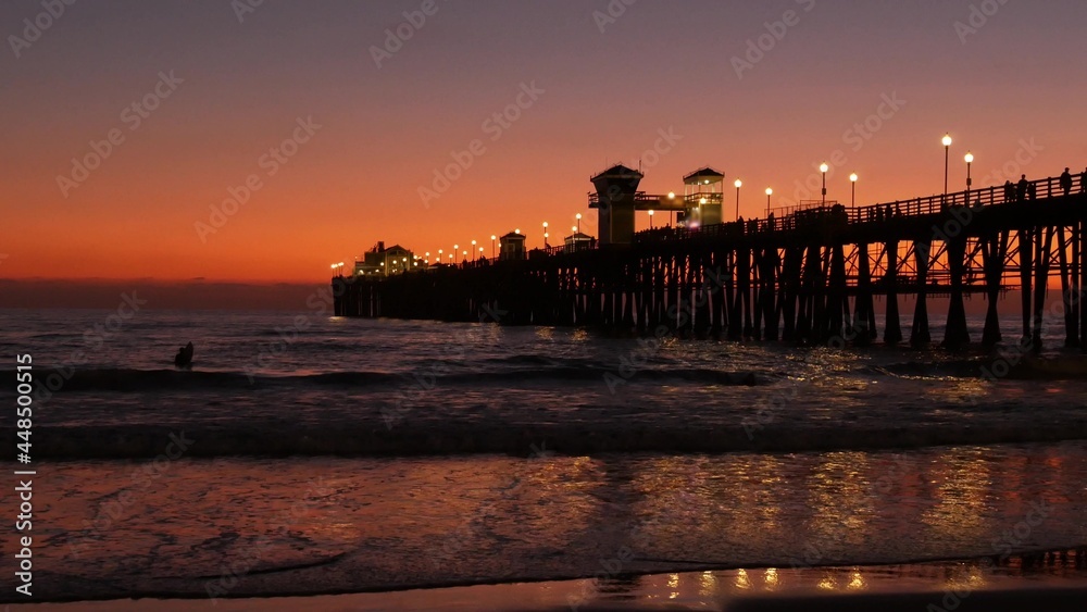 Wooden pier silhouette at sunset, California USA, Oceanside. Waterfront surfing resort, pacific ocean tropical beach. Summertime sea coastline vacations atmosphere. Surfers waiting for wave in water.