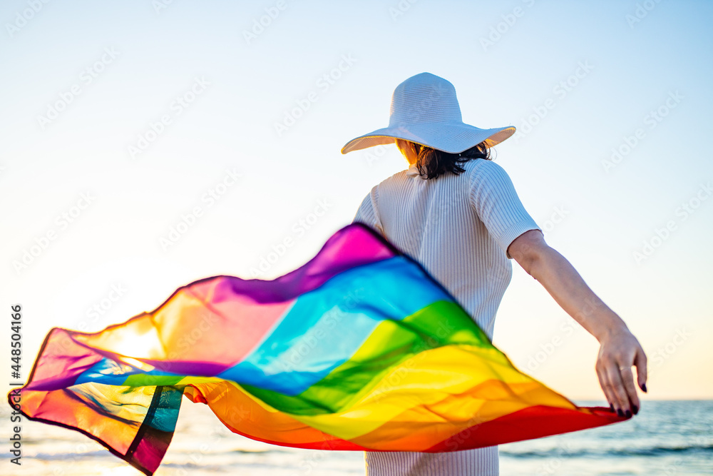 woman waving with rainbow flag and hat at sea beach at sunset