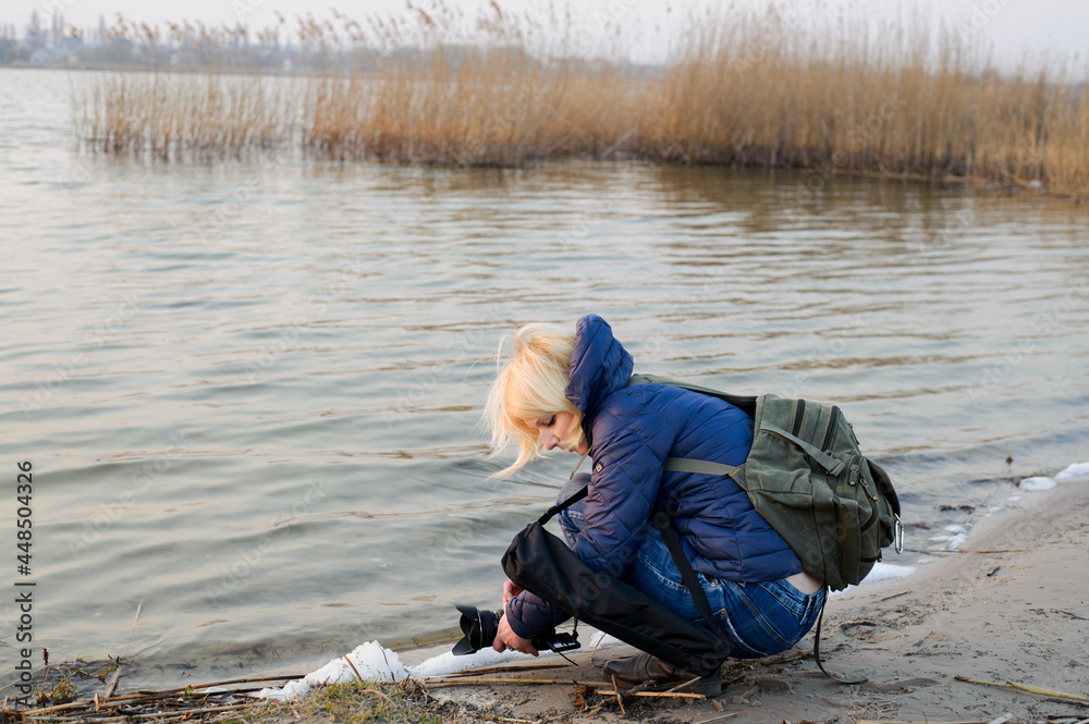 young girl photographer. photographer by the river, pictures outdoor, with camera and a backpack. blonde shoots water, Nature photography, autumn photo. editorial, April 2020, Ukraine, Kiev