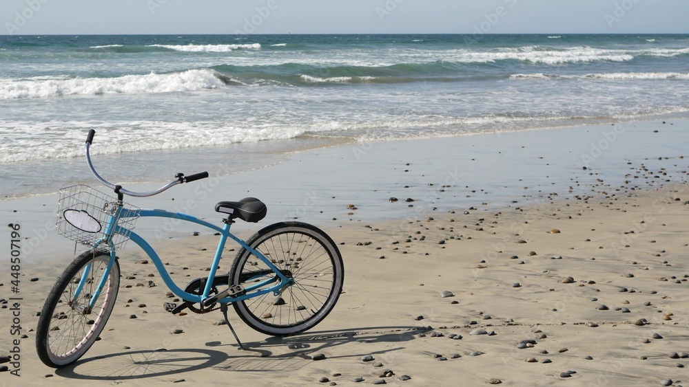 Fototapeta premium Blue bicycle, cruiser bike by ocean beach pacific coast, Oceanside California USA. Summertime vacations, sea shore. Vintage cycle on sand. Clear sky and water waves. Waterfront summer near Los Angeles