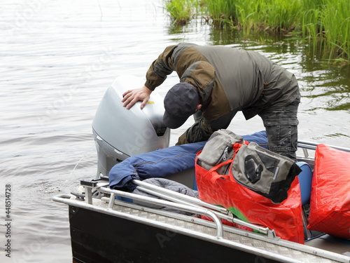 Boater man checks the hood of a 50 hp four stroke outboard motor on transom of boat near grass, repair and maintenance boat engine photo