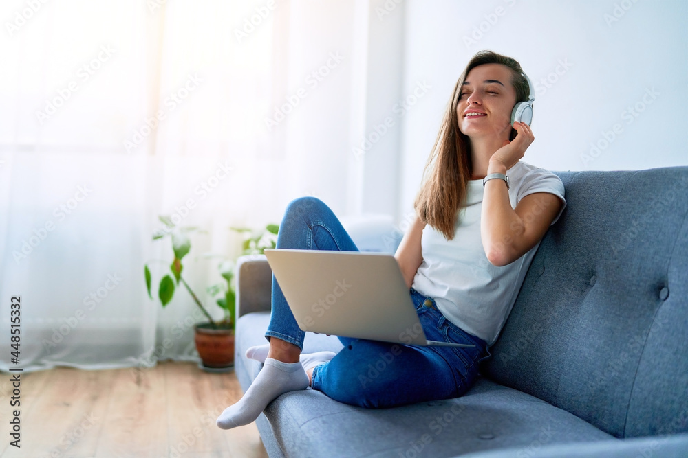 Young happy smiling joyful millennial girl music lover sitting on a sofa and using laptop with wireless headphones for enjoying listening music online at home Young happy smiling joyful millennial girl music lover sitting on a sofa and using laptop with wireless headphones for enjoying listening music online at home