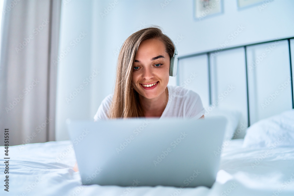 Young happy smiling joyful millennial girl lying on a bed and using laptop with wireless headphones for watching video online, learning language courses, social networking and video calling at home Young happy smiling joyful millennial girl lying on a bed and using laptop with wireless headphones for watching video online, learning language courses, social networking and video calling at home