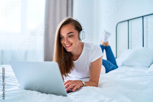 Young happy smiling joyful millennial girl lying on a bed and using laptop with wireless headphones for watching video online, learning language courses, social networking and video calling at home