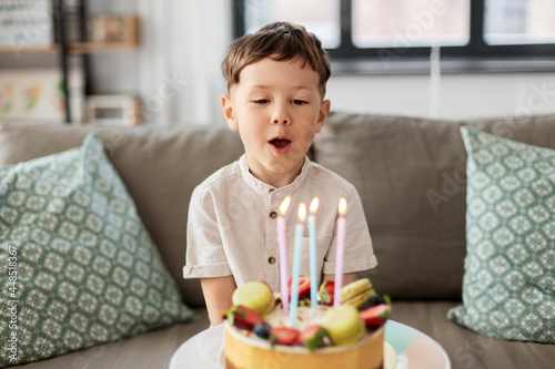 holidays, celebration and people concept - happy little boy blowing candles on birthday cake at home party