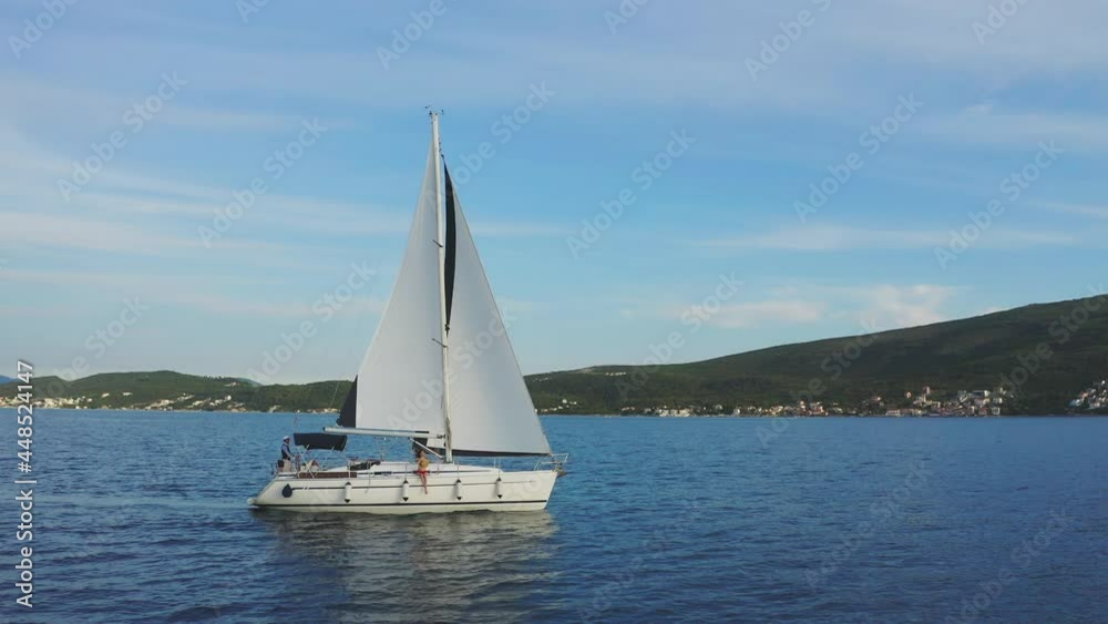 Side tracking of yacht sailing in sea bay with mountains in background. Woman in swimsuit sitting on side deck and swinging legs in air.