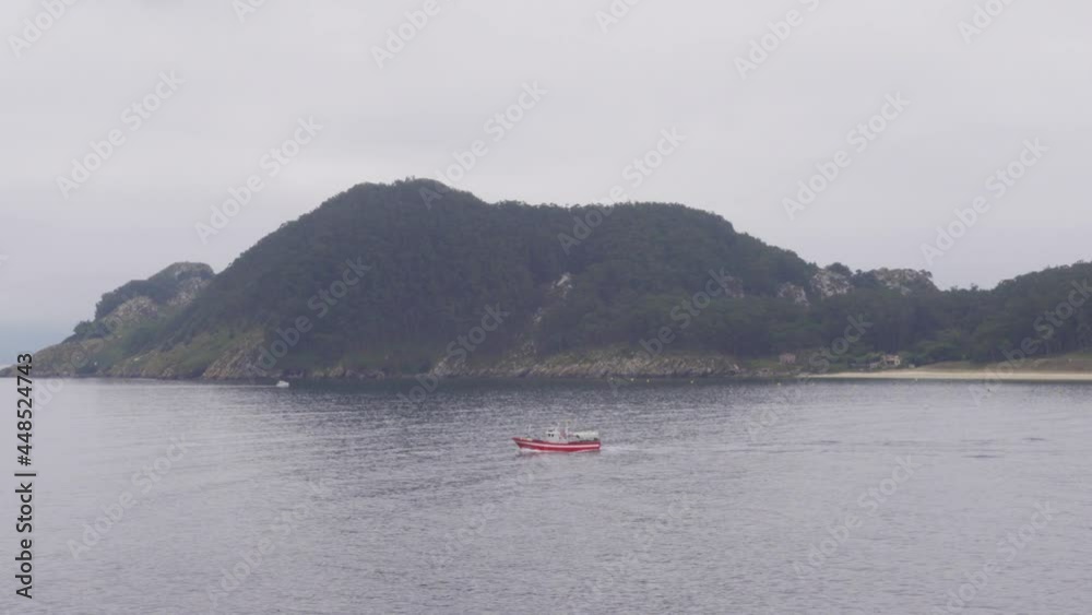 A Red Boat Passing by A Moody and Mysterious Uninhabited Island in CIes Islands Chain, Spain A Red Boat Passing by A Moody and Mysterious Uninhabited Island in CIes Islands Chain, Spain