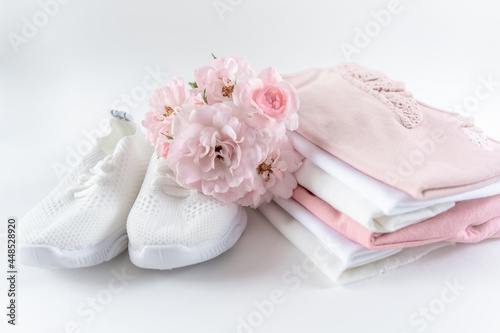 A stack of pink baby clothes and white sneakers. Toddler clothes set on white background with copy space decorated with pink flowers