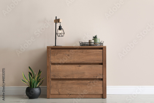 Wooden chest of drawers with houseplants and lamp near beige wall in room photo