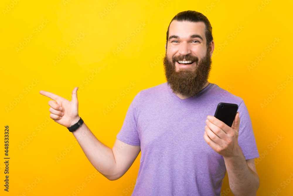 A cheerful bearded man is smiling at the camera holding his phone and pointing away at a free space A cheerful bearded man is smiling at the camera holding his phone and pointing away at a free space