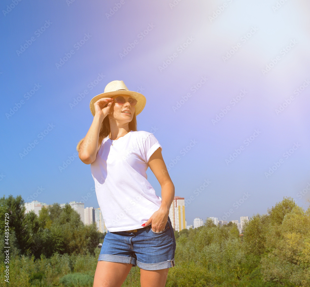Young woman in a hat and sunglasses against a background of green foliage, houses and blue sky. The sun shines brightly