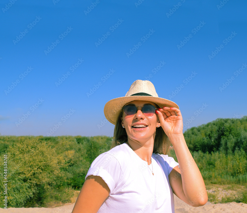 Portrait of a young cheerful woman in a hat and sunglasses on a background of a forest green and a blue sky