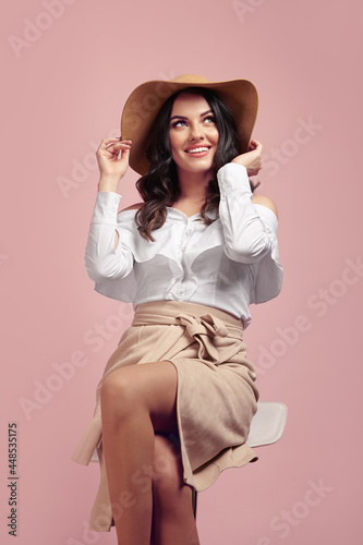 Attractive caucasian woman dressed in a beige hat and white shirt sits on high chair over pink isolated background  smiles happily  looks above the camera  wears a light brown skirt.
