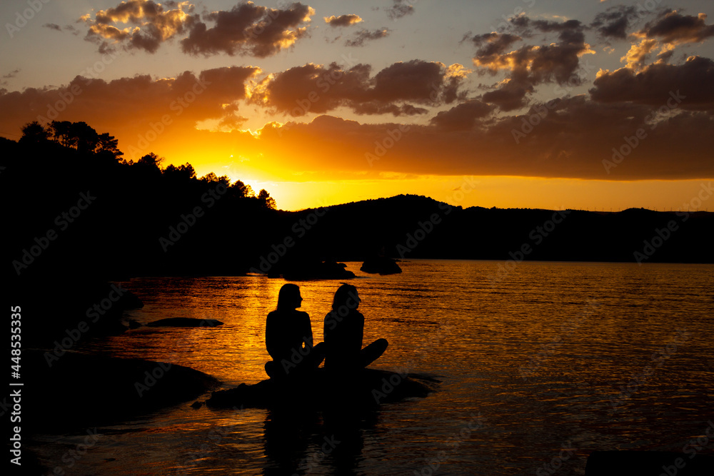Black silhouette of two friends sitting on a lake in the golden sunset. Copy space.