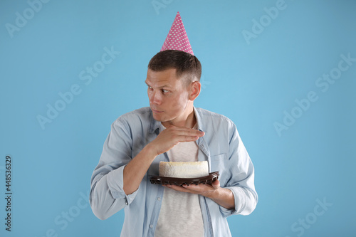 Greedy man with party hat hiding birthday cake on turquoise background photo