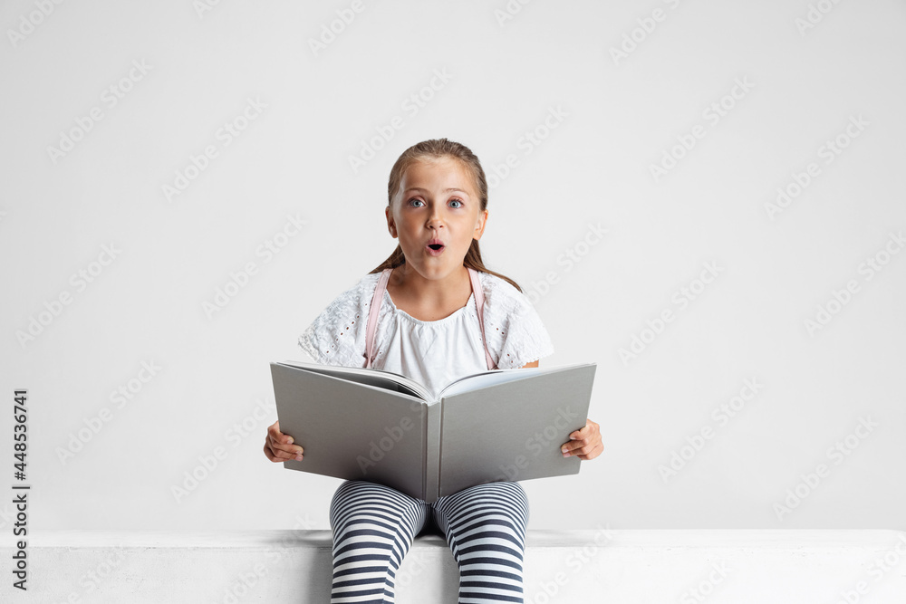 Portrait of cute Caucasian girl, teen sitting and reading big book, magazine isolated on gray studio background. Childhood, education, emotion concept. Portrait of cute Caucasian girl, teen sitting and reading big book, magazine isolated on gray studio background. Childhood, education, emotion concept.