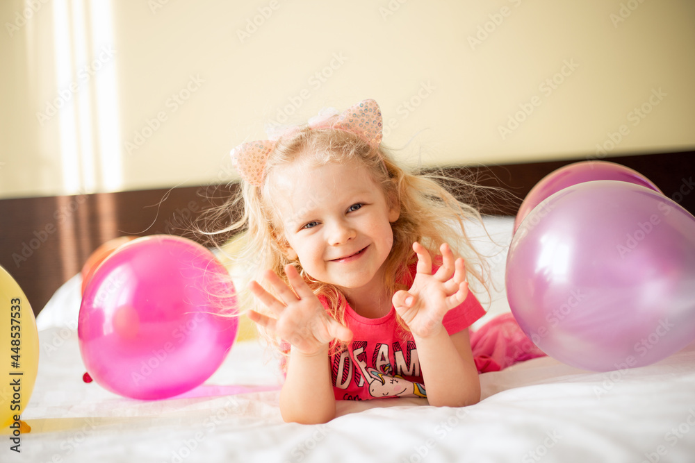Little cute caucasian sly baby girl lying on the bed with balloons in Children's room makes a wish and waiting friends. Happy Birthday concept Little cute caucasian sly baby girl lying on the bed with balloons in Children's room makes a wish and waiting friends. Happy Birthday concept