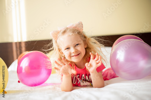 Little cute caucasian sly baby girl lying on the bed with balloons in Children's room makes a wish and waiting friends. Happy Birthday concept