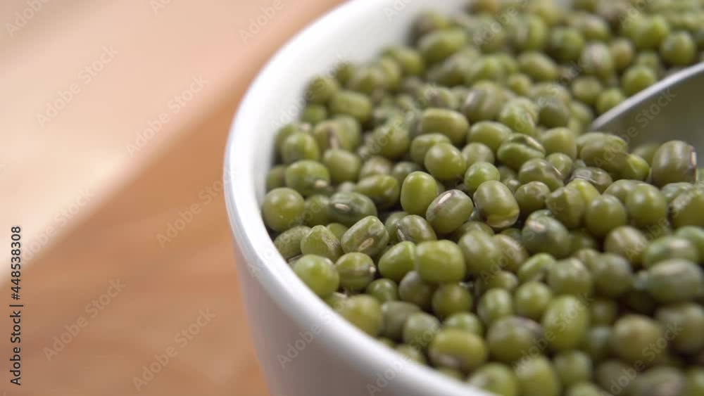 Full bowl of green mung beans on a wooden surface. Stir uncooked dry legumes with a spoon. Macro shot. Vigna radiata Full bowl of green mung beans on a wooden surface. Stir uncooked dry legumes with a spoon. Macro shot. Vigna radiata