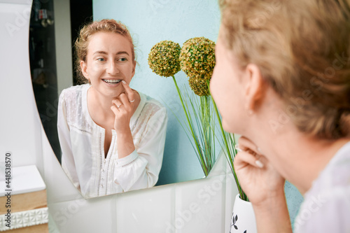 Woman with orthodontic brackets on teeth looking in the mirror. Charming woman in white elegant shirt checking braces on teeth in bathroom. Concept of dentistry, stomatology and orthodontic treatment. photo