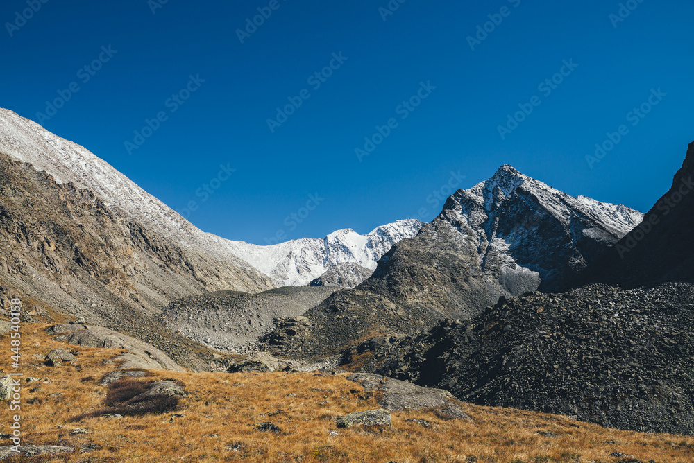 Autumn landscape with highland valley among snow-covered mountain range and pointy peak under blue sky. Atmospheric mountain scenery with sunlit orange valley in autumn colors among rocky mountains.