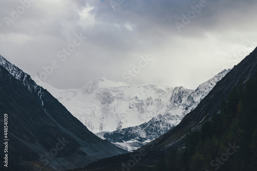 Atmospheric landscape with high snowy mountain wall and glacier in valley among dark silhouettes of rocks under cloudy sky. Dramatic view to snow-covered pinnacle. White snow mountains and black rocks