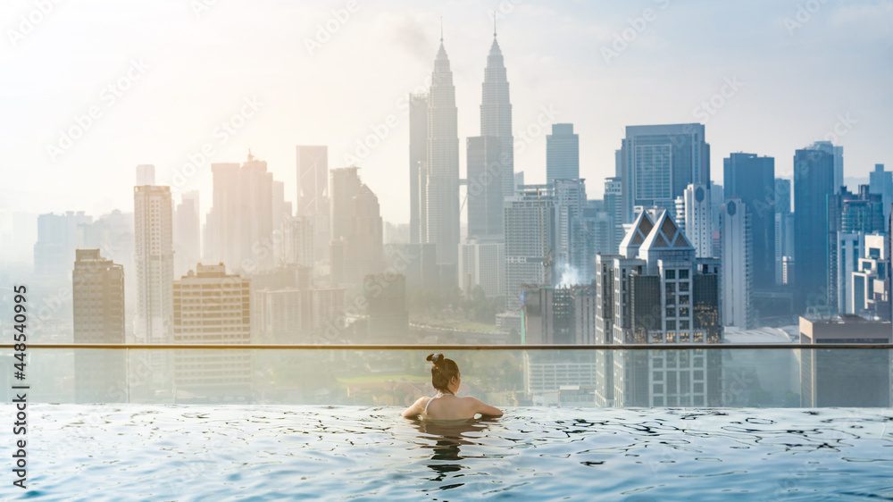Fototapeta premium young Asian woman enjoying in hotel roof top swimming pool, city view with blue sky for vacation travel, lifestyle of beautiful tourist girl with luxury outdoor skyscraper, Kuala Lumpur, Malaysia