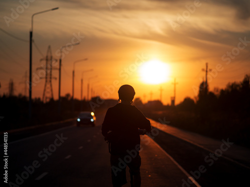 Silhouette of a young man on an electric scooter  a straight road ahead towards the sunset