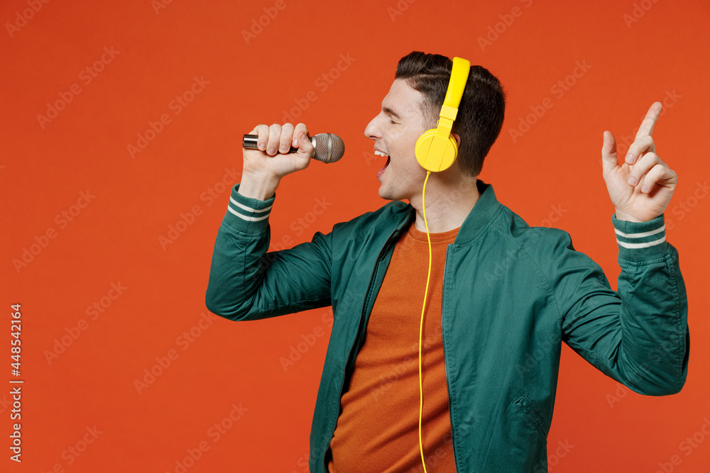 Cheerful satisfied young brunet man 20s wears red t-shirt green jacket listen to yellow headset headphones keep eyes closed singing in microphone isolated on plain orange background studio portrait