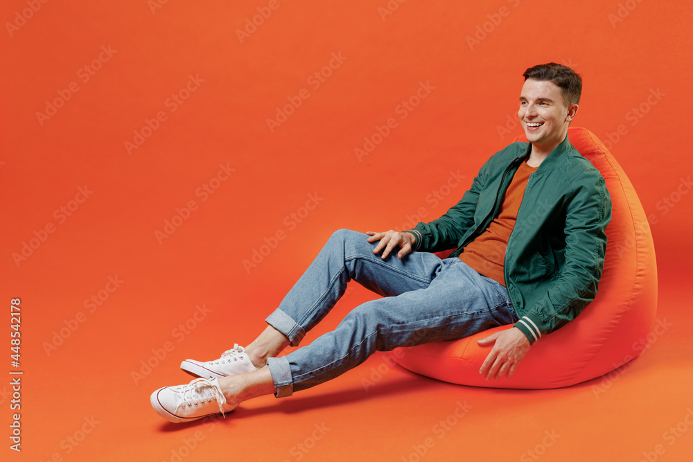 Full size body length happy young brunet man 20s wears red t-shirt green jacket sit in bag chair look aside away isolated on plain orange background studio portrait. People emotions lifestyle concept. Full size body length happy young brunet man 20s wears red t-shirt green jacket sit in bag chair look aside away isolated on plain orange background studio portrait. People emotions lifestyle concept.