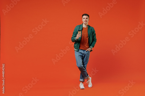 Full size body length smiling happy young brunet man 20s wears red t-shirt green jacket hold takeaway delivery craft paper brown cup coffee to go isolated on plain orange background studio portrait