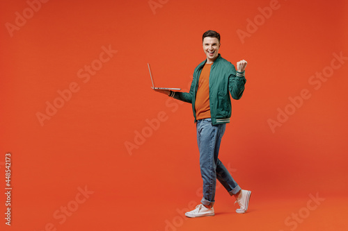 Full size body length vivid happy young brunet man 20s wears red t-shirt green jacket hold use work on laptop pc clenching fists say yes computer isolated on plain orange background studio portrait
