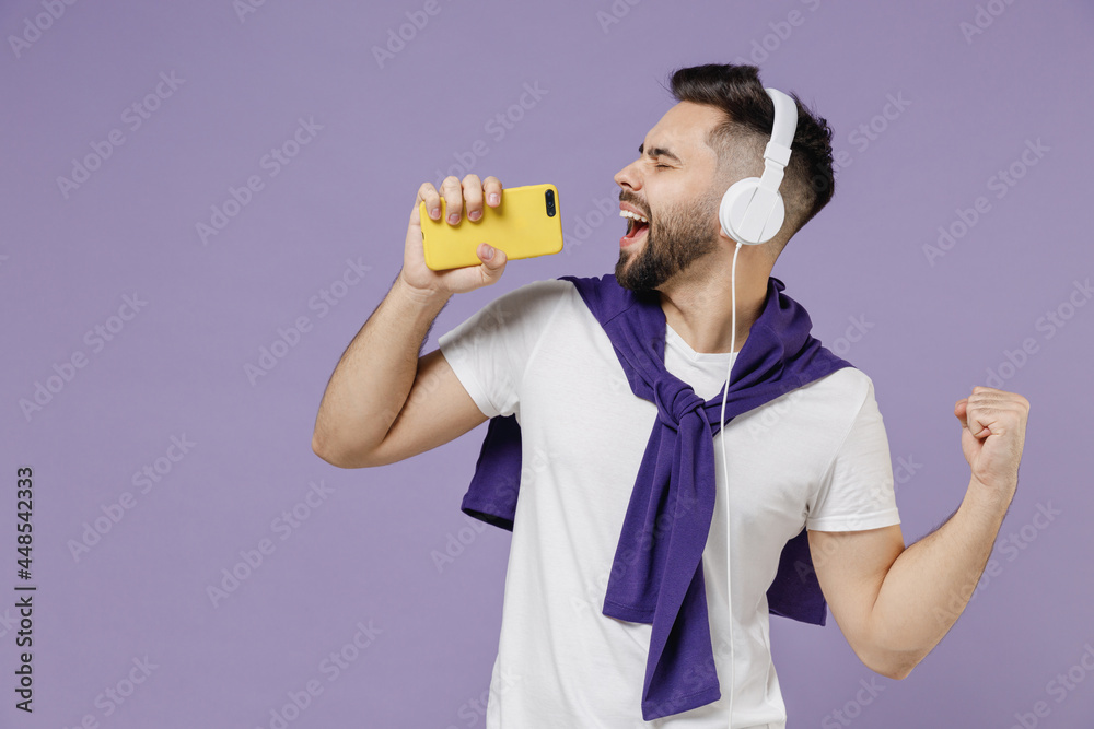 Side view profile young brunet man 20s wears white t-shirt purple shirt listen to music in headphones keep eyes closed sing in mobile cell phone isolated on pastel violet background studio portrait Side view profile young brunet man 20s wears white t-shirt purple shirt listen to music in headphones keep eyes closed sing in mobile cell phone isolated on pastel violet background studio portrait